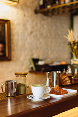 March 13, 2018, Belarus, Minsk. Vertical photo of a white illy coffee cup with matcha cappucсino and a plate with a croisant on a wooden bar counter.