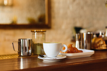 Obraz premium March 13, 2018, Belarus, Minsk. Horizontal photo of a white illy coffee cup with matcha cappucсino and a plate with a croisant on a wooden bar counter.