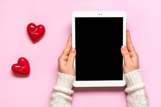 female hands holding digital tablet for chooses gifts, makes purchase, two red hearts on pink table Top view Flat lay Holiday shopping list, Happy Valentine's day, party, online shop concept Mockup