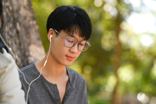Photo of young asian man reading a book and listening to music in earphones with his girlfriend
