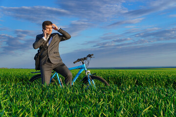 a businessman rides a bicycle on a green grassy field, dressed in a business suit, he makes a phone call, beautiful nature in spring, freelance business concept