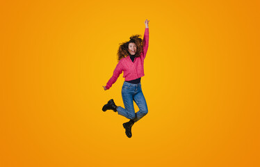 Fototapeta na wymiar Full length portrait of a cheerful young curly-haired woman celebrating success while jumping isolated over yellow background. Happiness, freedom, power, motion