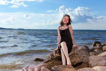 One Winsome Positive Young Happy Smiling Caucasian Brunette Girl in Black Dress With Sincere Smile Resting At Seashore Stones During Sunny Day Outdoors.
