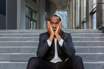 Fototapeta premium Upset african american man sitting on stairs outside office building, businessman bankrupt in despair, mature boss depressed in business suit outdoors.