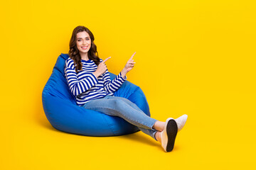 Full length portrait of cheerful nice person sit comfy bag indicate fingers empty space isolated on yellow color background