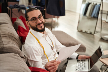 Cheerful fashion designer with laptop and sketches in his workshop