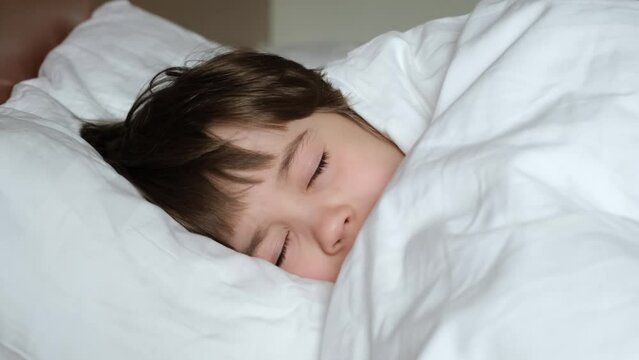 One happy boy in bed under blanket tries to sleep. The child looks at camera then closes his eyes and fall asleep. Daytime relax. White linens. Dreaming time concept. Soft pillow and warm blanket.