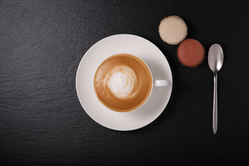 White cup with cappuccino on a dark background flat lay. breakfast on a black background. Coffee with macaroon and a spoon on a black background.