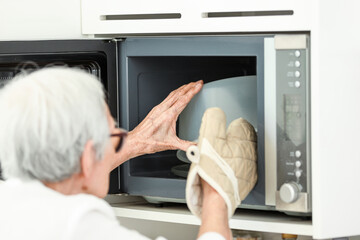 Asian senior woman wearing oven glove while taking food out of microwave oven,prevent damage and injury from heat accident,safety at home,hot bowl from cooking,old elderly heating food in kitchen.