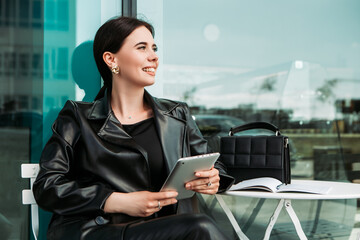 Fototapeta premium Pretty brunette caucasian woman dressed leather suit with shoulder bag sitting outside cafe street hold tablet pc. Businesswoman office clerk. Smiling white teeth lady. Professional business owner 