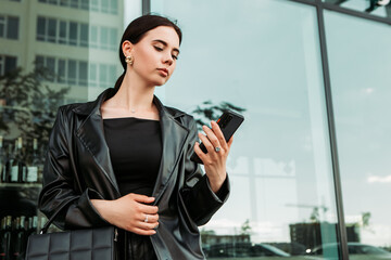 Pretty brunette caucasian woman dressed leather suit with shoulder bag walking outside hold mobile phone, order taxi, ride sharing, typing message texting chatting, search information on the Internet
