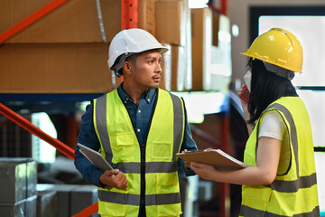 Two warehouse coworkers talking to each other while standing between rows of tall shelves full of packed boxes and goods