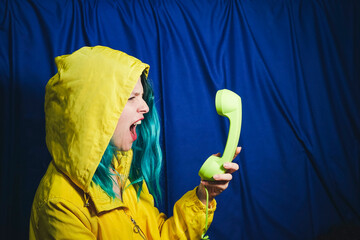 Woman in yellow hooded shirt shouting into telephone in front of blue backdrop