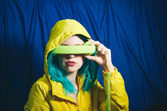 Woman in hooded shirt covering eyes with telephone receiver in front of blue backdrop