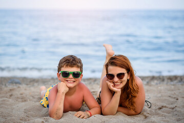  young mother and her son in sunglasses lying on the beach sand near the sea and smiling..