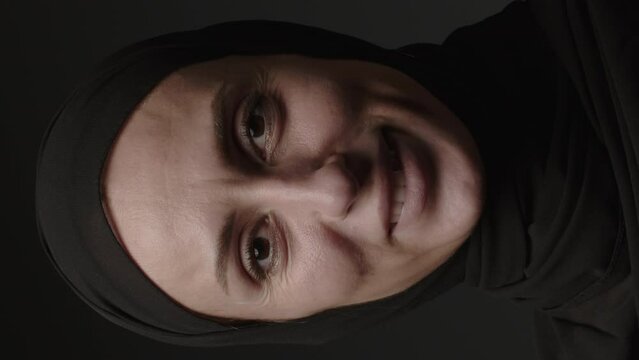 Vertical video of close-up portrait of a Muslim woman with beautiful eyes in a hijab on a dark background. A girl in a black headscarf is looking at the camera. Arab women life concept