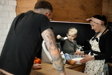Happy family from three people cook dinner. Woman hold deep glass bowl with flour, little boy take spoonful of flour.