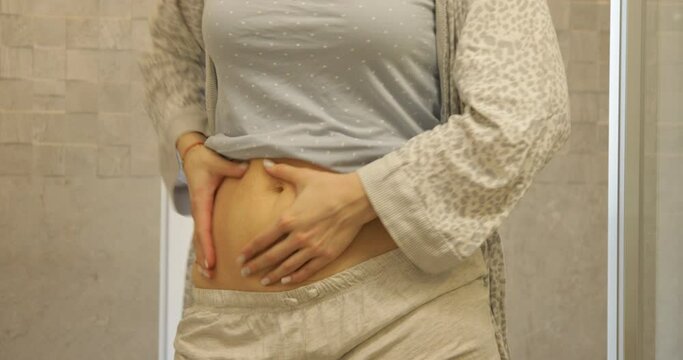 Unrecognizable woman wearing grey home wear, standing in bathroom, doing massage on naked belly, shaking, lifting.