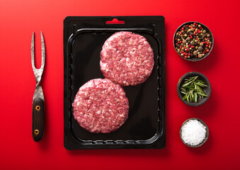 Beef burgers in vacuum tray with fork and salt and pepper on red background.