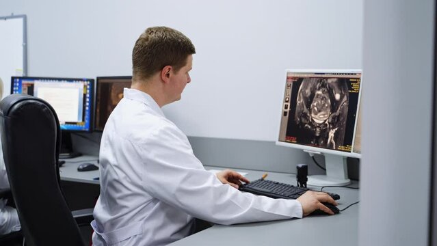 Male lab technician wearing white robe sitting at the table. Professional checking up different scans on the computer screen. MRI shots analysis.