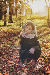 Pensive little girl model in brown coat sitting in autumn park outdoor, looking away. Pretty young lady walking in fall forest leisure. Childhood youth style concept. Copy text space for advertising