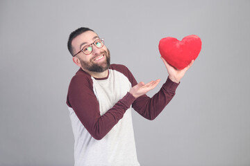 Young man holding a small red heart shaped pillow while looking at camera - 565302964