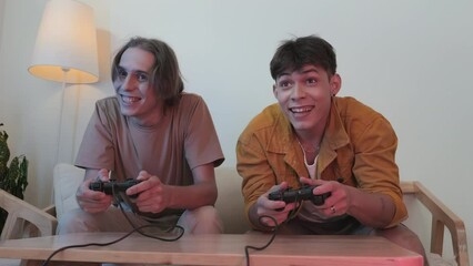 Two Cheerful boys in their early 20s having fun at home playing video games together with controllers on couch