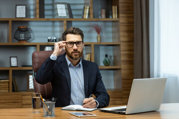 Fototapeta na wymiar Serious thinking boss inside the office portrait of a mature businessman with a beard and glasses, the man is looking at the camera with concentration, working with documents on a laptop at the table