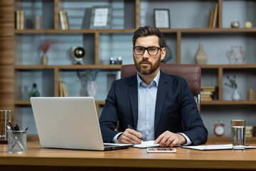 Serious thinking boss inside the office portrait of a mature businessman with a beard and glasses, the man is looking at the camera with concentration, working with documents on a laptop at the table