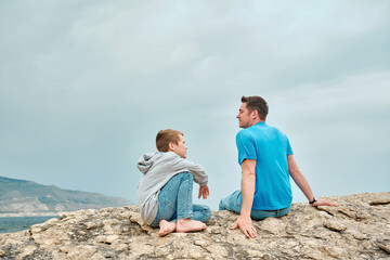 Happy family, father and son bonding, sitting on stone by the sea looking at view enjoying summer vacation. Togetherness Friendly concept