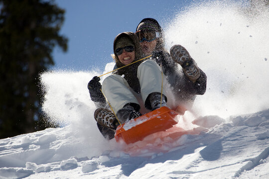 Two girls barrel down a slope on a plastic sled. Snow sprays from the sled as they continue to gather speed.