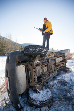 A hunter holds his rifel while standing on his flipped truck after an accident.