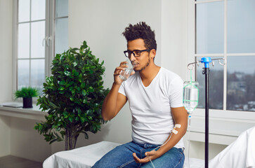 Portrait of a sick young african american man in the glasses sitting in a white medical ward under...