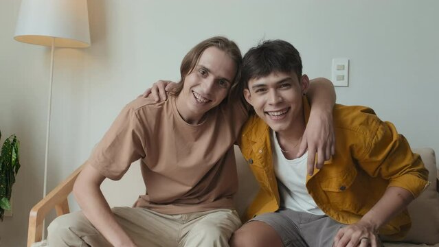 Slowmo portrait of two buddies in their early 20s sitting together on couch indoors pointing and smiling at camera