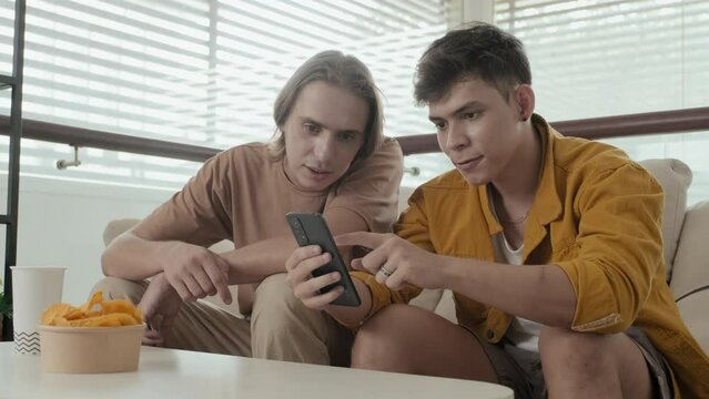 Two Caucasian Male Friends Looking At Smartphone And Chatting While Hanging Out At Home Together