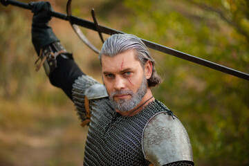 An aged male warrior in chain mail with gray hair and a scar on his face. Knight in armor holds a two-handed sword.