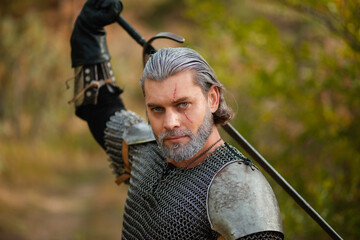An aged male warrior in chain mail with gray hair and a scar on his face. Knight in armor holds a two-handed sword.