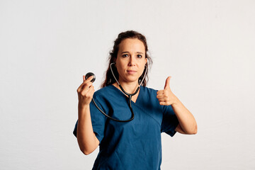 A doctor holding her stethoscope with one hand and showing thumb up with the other hand while looking at camera in a studio - 565320748