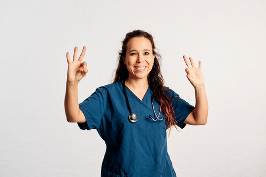 A doctor standing smiling and looking at camera while she performs ok gestures with both hands in a studio