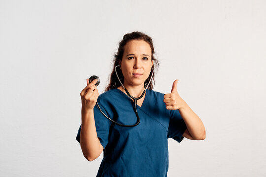 A doctor holding her stethoscope with one hand and showing thumb up with the other hand while looking at camera in a studio
