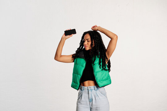 A young woman dancing as her arms raised while holding her cell phone in a studio shot