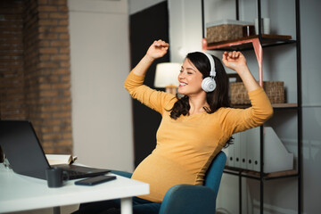 Beautiful pregnant listening the music while working on laptop. Young businesswoman working in her office.