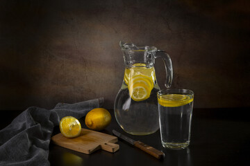 Water with lemon in a glass jug and in a glass on a dark background. Useful drink for health