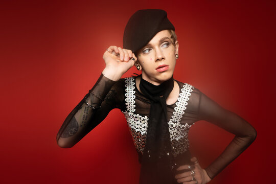 fashionable queer model adjusting black beret while standing with hand on hip and looking away on red background.