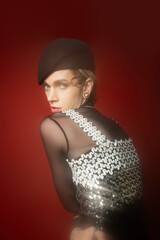 young nonbinary model in elegant top with sequins looking at camera on dark red background.