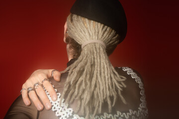 back view of queer person with dreadlocks and silver rings holding hand on shoulder on dark red background.