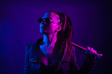 A young woman with dreadlocks in the dark in a club in black glasses holds a painless bat in her hands. Girl with afro-braids at night in neon color.