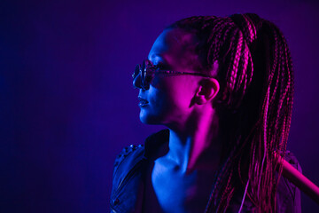 A young woman with dreadlocks in the dark in a club in black glasses holds a painless bat in her hands. Girl with afro-braids at night in neon color.