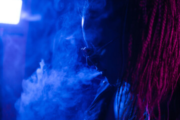 A young woman with dreadlocks smokes a cigarette in a club. Wearing black glasses, he holds a painless bat in his hands. Girl with afro pigtails in the smoke at night in neon color. Scoundrel