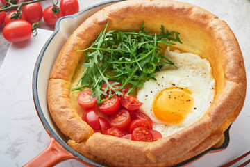Dutch baby pancake. Fresh homemade Dutch Baby pancake with fried egg, tomato and green arugula in reed cast-iron pan on light gray concrete rustic table. Flat lay, close up.